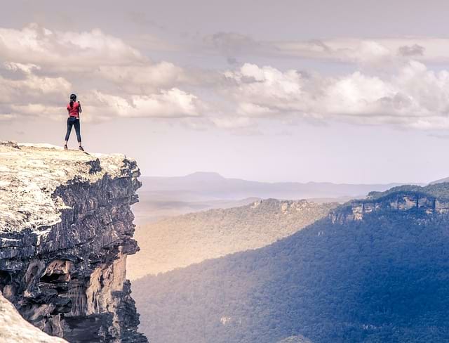 Photo of a woman standing at the top of a mountain, watching over a cliff at the mountains on the other side of the valley. Like this hiker, as participants reach the end of the Big Picture Event Storming, a whole new world of possibilities open to them!